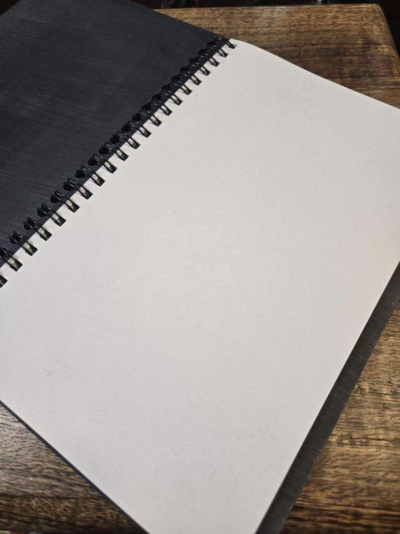 May include: An open spiral-bound notebook with a black cover and blank white pages. The notebook is lying on a wooden surface, with the pages open to reveal the clean, unlined paper. The spiral binding is visible along the left edge.