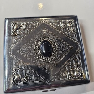 Pode incluir: Um porta-cigarros de metal prateado com um design ornamentado. A caixa apresenta um padrão central em forma de diamante com uma pedra oval preta e detalhes florais e de rolagem decorativos. Os cantos têm detalhes florais estilizados.