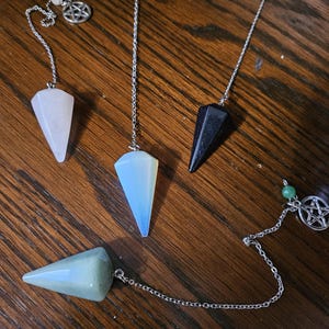 May include: Four crystal pendulums with silver chains and pentagram charms. The pendulums are made of clear, white, blue, and green crystals.