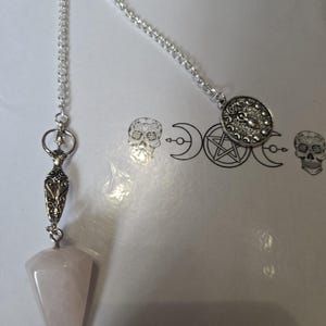 May include: A rose quartz pendulum with a silver chain and decorative accents. The pendulum has a pointed, faceted shape. The chain is attached to a decorative silver piece and a circular charm. The background features occult symbols.