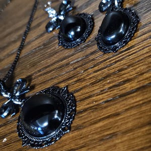 Könnte beinhalten: Ein schwarzes Halsketten- und Ohrring-Set mit einem schwarzen Kameen-Anhänger und passenden Ohrringen. Anhänger und Ohrringe haben einen schwarzen ovalen Stein, der von einem filigranen schwarzen Design umgeben ist. Jeder Ohrring hat eine schwarze Schleifenverzierung.
