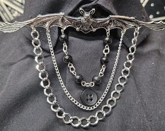May include: A silver-toned metal brooch featuring a bat with outstretched wings. The brooch is adorned with multiple dangling silver chains, a strand of small black beads, and a single black four-hole button suspended from the center chains.