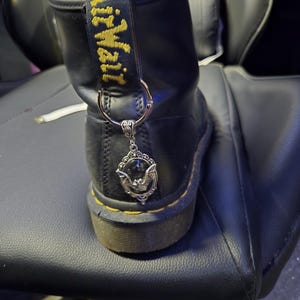 May include: A black leather boot with a yellow AirWair heel loop and a silver-toned metal charm attached to the back. The charm features an ornate oval frame with a detailed bat figure in the center, suspended from a circular metal ring.