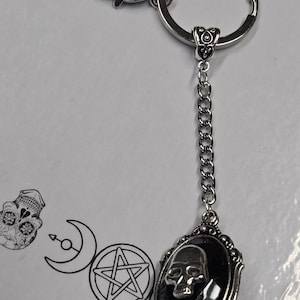 May include: Silver keychain featuring a skull pendant. The oval pendant has a black backdrop and a detailed skull design. The keychain includes a chain and a heart-shaped clasp. The background has a skull illustration and occult symbols.
