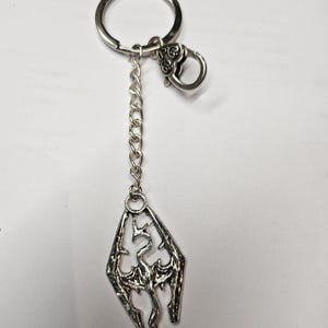 May include: A silver-toned metal keychain featuring a circular split ring, a small lobster clasp, and a short chain. The pendant is a diamond-shaped emblem with an intricate, stylized dragon cutout design in a weathered, metallic finish.