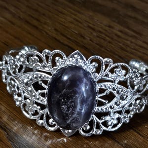 May include: A silver-toned cuff bracelet featuring an intricate filigree design with floral motifs and a central oval-shaped dark purple stone with visible white marbling patterns throughout the surface of the gemstone.