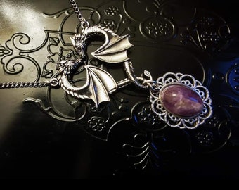 Puede incluir: Un collar de metal plateado que presenta dos figuras de dragón con las alas extendidas frente a frente, conectadas a una piedra preciosa ovalada de color púrpura engastada en un marco de metal de filigrana con un borde festoneado.
