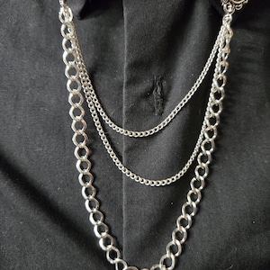 May include: A silver-toned collar chain accessory featuring two oval-shaped black cabochon brooches with metallic accents. The brooches are connected by three layered silver chains of varying thicknesses that drape across a black button-down shirt.