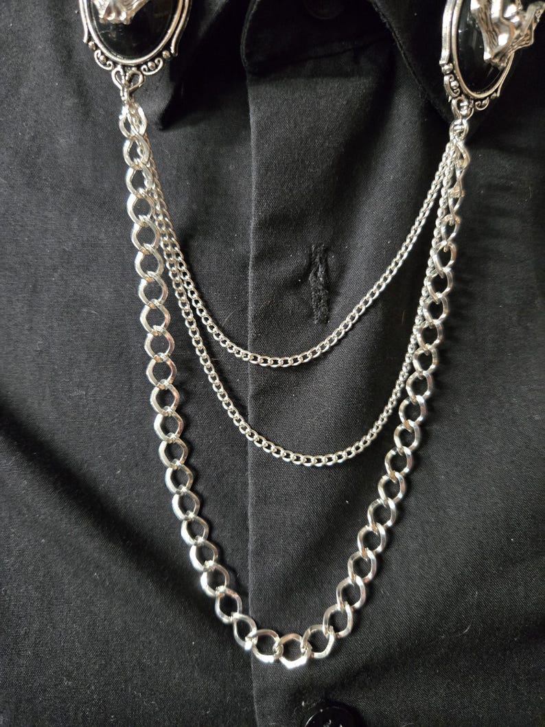 May include: A silver-toned collar chain accessory featuring two oval-shaped black cabochon brooches with metallic accents. The brooches are connected by three layered silver chains of varying thicknesses that drape across a black button-down shirt.