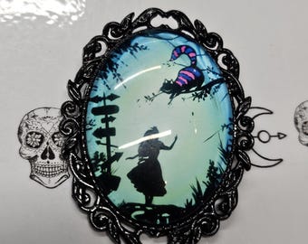 May include: An oval brooch featuring a black ornate metal frame surrounding a glass cabochon. The image inside depicts a silhouette of a person standing near a signpost, with a striped cat perched on a tree branch against a light blue background.