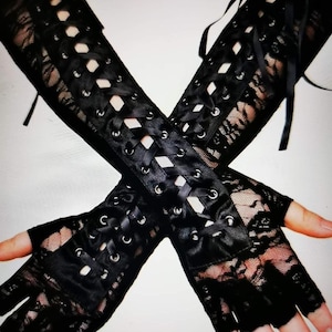 May include: A pair of black fingerless gloves featuring intricate floral lace fabric and a central vertical panel with silver-toned metal eyelets. The gloves are secured with a crisscross black satin ribbon lacing system that extends along the forearm.