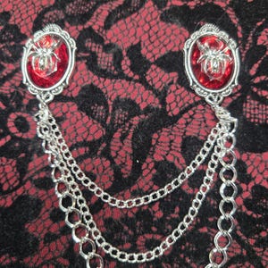 May include: A silver-toned brooch featuring two oval settings with faceted red glass cabochons, each centered with a metallic spider emblem. The two settings are connected by three draped silver-toned metal chains of varying link sizes.