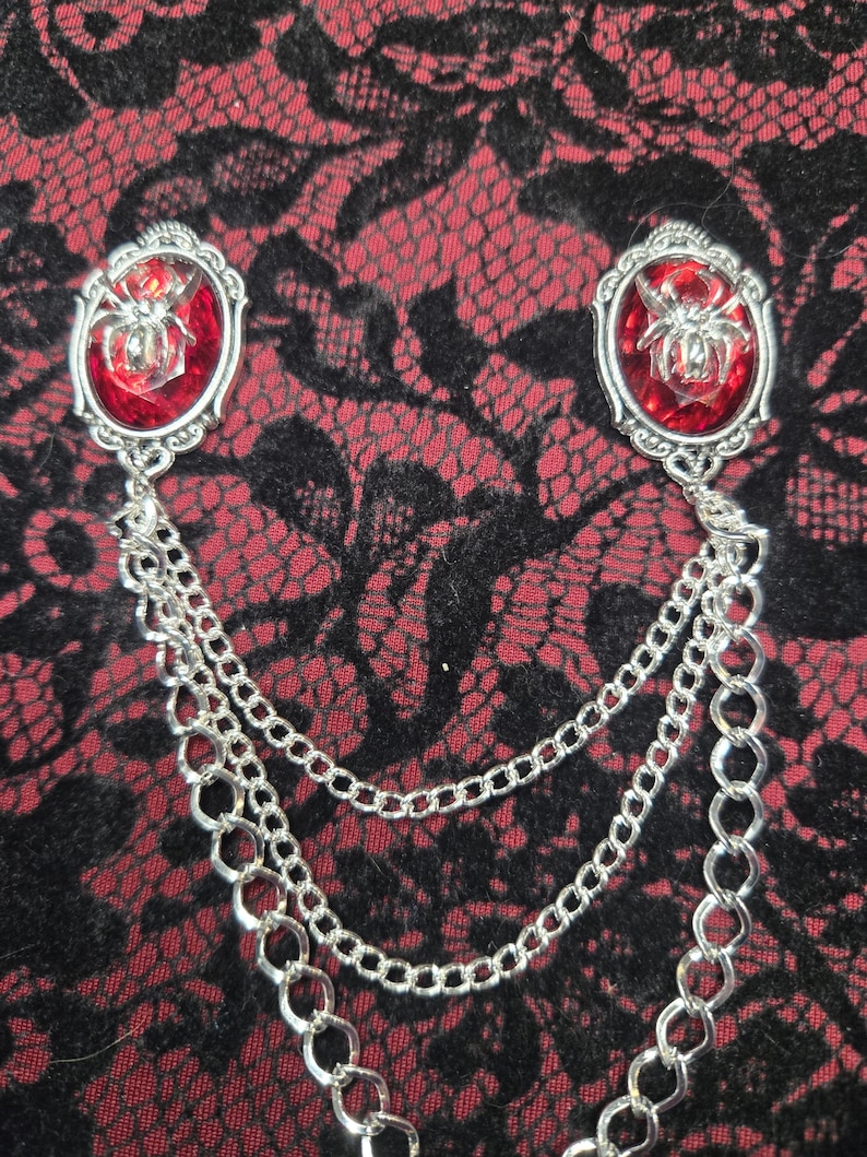 May include: A silver-toned brooch featuring two oval settings with faceted red glass cabochons, each centered with a metallic spider emblem. The two settings are connected by three draped silver-toned metal chains of varying link sizes.