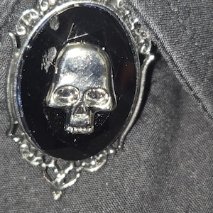 May include: An oval brooch featuring a silver-toned skull emblem centred on a smooth black stone. The piece is encased in an ornate, filigree-style silver metal frame with decorative scrollwork details along the edges of the oval setting.