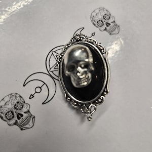 May include: A silver-toned oval brooch featuring a three-dimensional metallic skull centered on a smooth black oval inlay. The frame has an ornate, beaded edge with decorative metalwork. The brooch is placed over a white surface with line art.