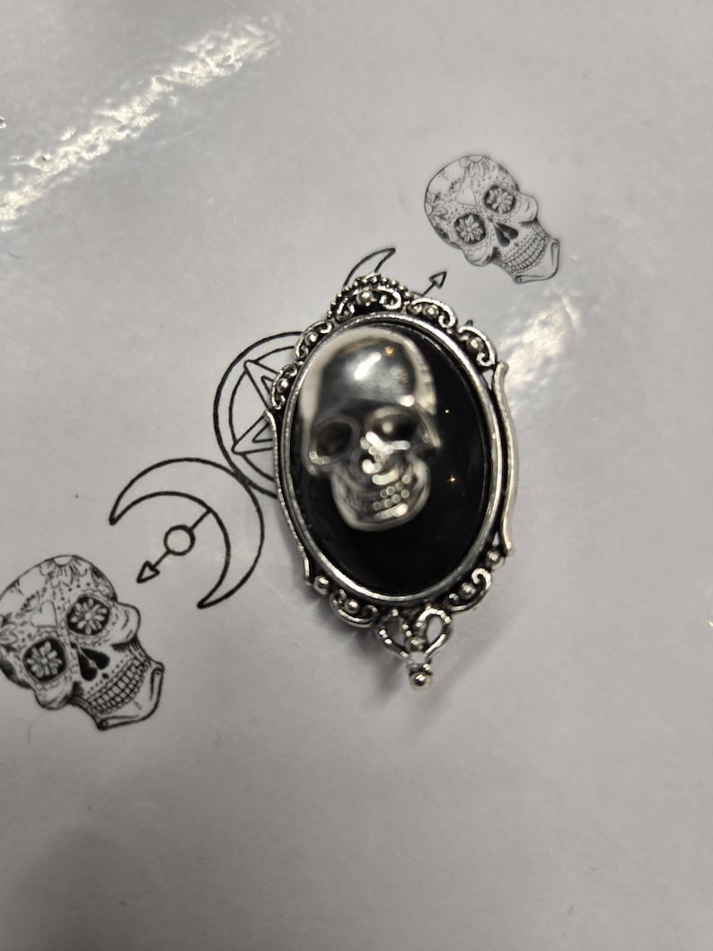 May include: A silver-toned oval brooch featuring a three-dimensional metallic skull centered on a smooth black oval inlay. The frame has an ornate, beaded edge with decorative metalwork. The brooch is placed over a white surface with line art.