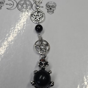 May include: A silver-toned necklace featuring a pentagram pendant, black beads, and a skull pendant with a black orb. The necklace has a chain and is likely made of metal. The design has a gothic or occult theme.