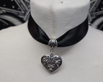 Może przedstawiać: Czarny naszyjnik typu choker z satynowej wstążki ze srebrzystą metalową zawieszką w kształcie serca. Medalion posiada wytłoczony szczegółowy wzór nietoperza na przedniej powierzchni i jest zawieszony na małym srebrzystym metalowym kółeczku.