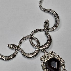 Puede incluir: Un collar de serpiente de plata con un colgante de gema negra. La serpiente está enroscada en forma de ocho y el colgante es de forma de diamante con una gema negra en el centro.