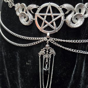 May include: A silver-toned waist chain featuring a central pentagram emblem flanked by two textured snake figures. The design includes multiple draped chain layers and a dangling filigree pendant with a small, dark, faceted stone drop element.