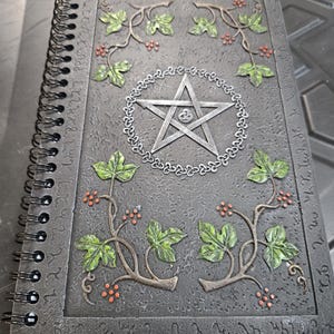 May include: A spiral-bound journal with a textured, dark gray cover. The cover features a raised design of a silver pentagram within a circle, surrounded by green leaves and red berries. The edges of the cover have an embossed pattern.