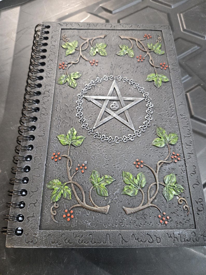 May include: A spiral-bound journal with a textured, dark gray cover. The cover features a raised design of a silver pentagram within a circle, surrounded by green leaves and red berries. The edges of the cover have an embossed pattern.