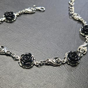 May include: A silver-toned metal bracelet featuring alternating black rose charms and small skull motifs. The chain links connect each floral element, and the piece includes a lobster claw clasp for closure, creating a cohesive and detailed accessory.