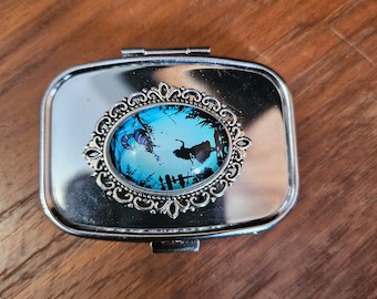 May include: A silver compact mirror with a round, blue and white glass cameo featuring a silhouette of a woman in a forest setting. The cameo is surrounded by an ornate silver frame.