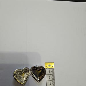 Pu&ograve; includere: Un medaglione argentato a forma di cuore aperto, che rivela un intricato motivo in rilievo sulla parte interna sinistra e una superficie liscia su quella destra. Un metro a nastro giallo posto accanto indica una larghezza di circa 3,8 cm.