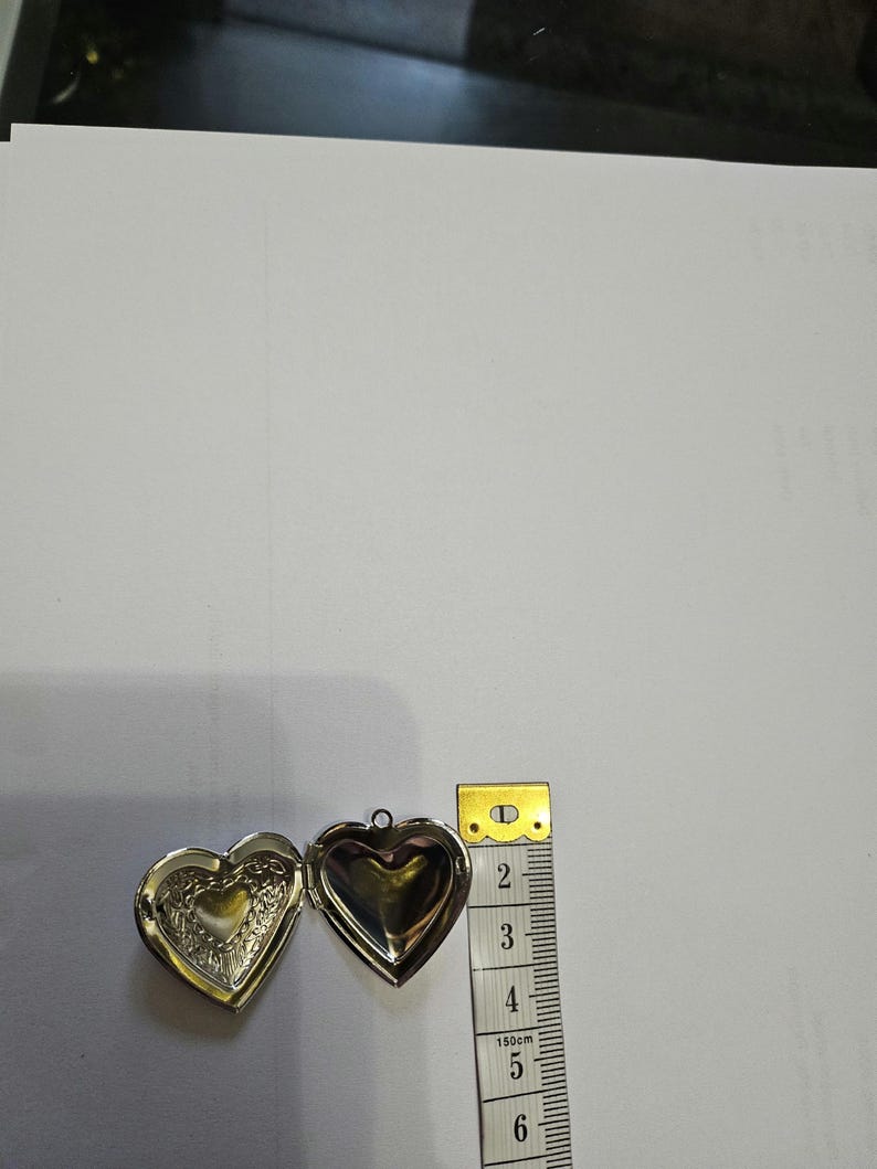 Pu&ograve; includere: Un medaglione argentato a forma di cuore aperto, che rivela un intricato motivo in rilievo sulla parte interna sinistra e una superficie liscia su quella destra. Un metro a nastro giallo posto accanto indica una larghezza di circa 3,8 cm.