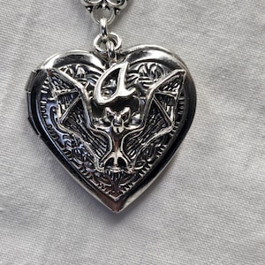 Puede incluir: Medallón de plata en forma de corazón con un diseño detallado de un murciélago con las alas extendidas. El medallón tiene una abertura con bisagra.