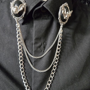 May include: A black collared shirt featuring two silver-toned oval brooches, each displaying a central black stone and a silver bat emblem. The brooches are connected by three draped silver chains of varying thicknesses that hang across the chest.