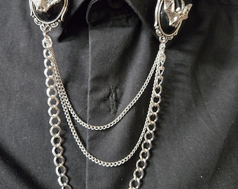 May include: A black collared shirt featuring two silver-toned oval brooches, each displaying a central black stone and a silver bat emblem. The brooches are connected by three draped silver chains of varying thicknesses that hang across the chest.