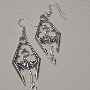 May include: A pair of silver-toned metal earrings featuring a diamond-shaped frame with an intricate dragon cutout design. Each earring hangs from a standard silver-coloured hook wire, showcasing a detailed mythical creature silhouette.