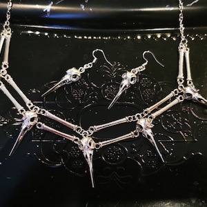 May include: A silver-toned jewelry set consisting of a necklace and a pair of earrings. The necklace features multiple bird skull pendants connected by thin metal bars and chain links. The matching earrings also feature bird skull pendants.
