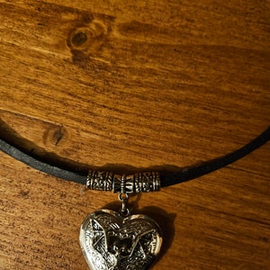 Pu&ograve; includere: Un ciondolo a forma di cuore color argento con un design a pipistrello in rilievo, sospeso a una collana con cordino nero. Il medaglione &egrave; fissato al cordino tramite una perlina decorativa in metallo color argento con motivi complessi.