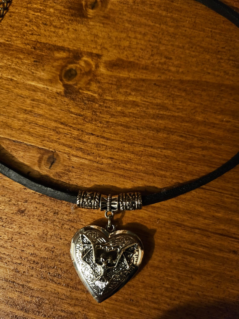 Pu&ograve; includere: Un ciondolo a forma di cuore color argento con un design a pipistrello in rilievo, sospeso a una collana con cordino nero. Il medaglione &egrave; fissato al cordino tramite una perlina decorativa in metallo color argento con motivi complessi.