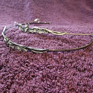 May include: A silver-toned metal headband featuring a detailed bat motif with outstretched wings. The bat is positioned along the thin, curved band, which has a smooth, polished finish. The design is symmetrical and metallic in appearance.
