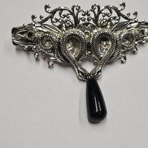 May include: A silver-toned metal hair clip featuring an intricate filigree design with two coiled snakes facing each other. A smooth, polished black teardrop-shaped stone hangs from the centre of the snakes, creating a decorative focal point.
