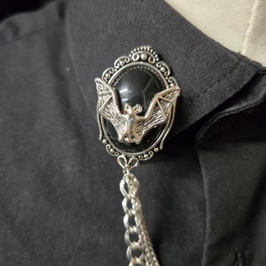 May include: A silver-toned brooch featuring an oval black cabochon set within an ornate frame. A sculpted silver bat is centered on the black stone. Two silver chains hang from the bottom of the brooch, which is pinned to a black collared shirt.