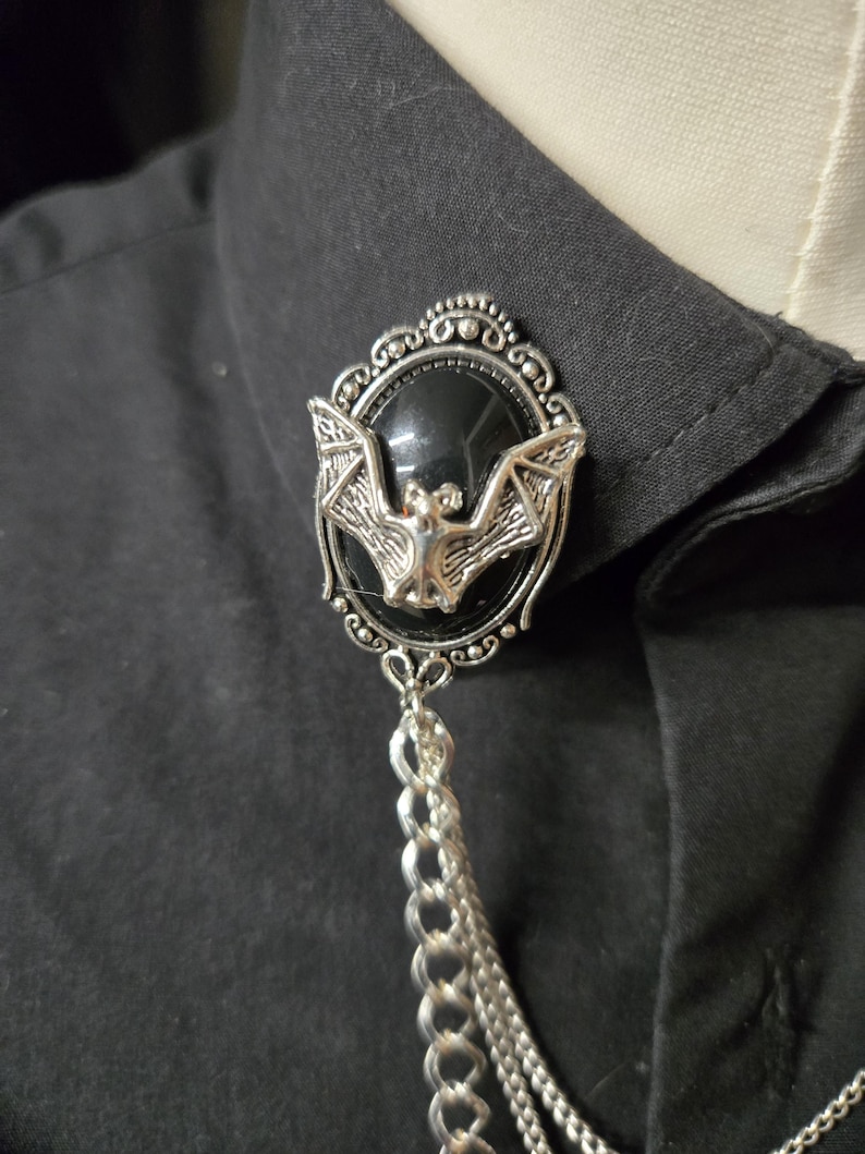 May include: A silver-toned brooch featuring an oval black cabochon set within an ornate frame. A sculpted silver bat is centered on the black stone. Two silver chains hang from the bottom of the brooch, which is pinned to a black collared shirt.