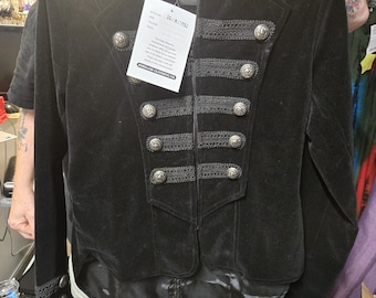 May include: A black velvet military-style jacket featuring a high collar, long sleeves, and a front closure adorned with five rows of decorative black braided trim and silver-toned metal buttons. A white product tag is attached to the collar.