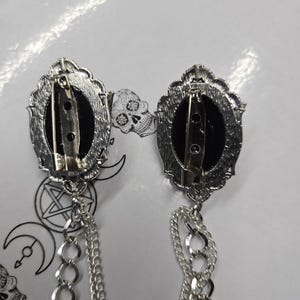 May include: Two silver-toned oval brooches with black centers, each featuring a pin backing and a decorative scalloped frame. Attached to the bottom of each brooch are two dangling silver-toned metal chains of varying link sizes.