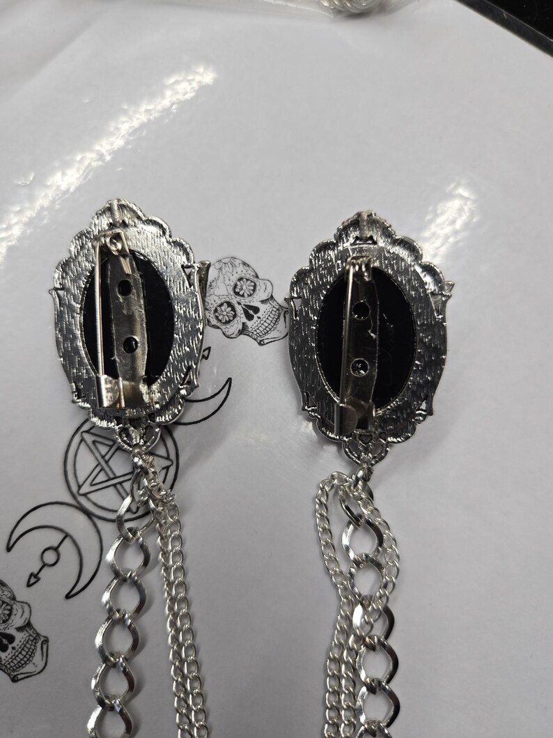 May include: Two silver-toned oval brooches with black centers, each featuring a pin backing and a decorative scalloped frame. Attached to the bottom of each brooch are two dangling silver-toned metal chains of varying link sizes.