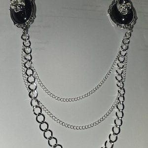 May include: A silver-toned brooch featuring two oval black cabochons, each topped with a small metallic skull. The two pieces are connected by three draped silver chains of varying thicknesses, creating a layered, dangling effect.