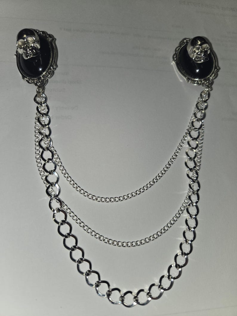 May include: A silver-toned brooch featuring two oval black cabochons, each topped with a small metallic skull. The two pieces are connected by three draped silver chains of varying thicknesses, creating a layered, dangling effect.