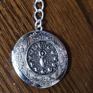 May include: Silver locket necklace with a detailed design featuring a stylized female figure and a crescent moon pattern.