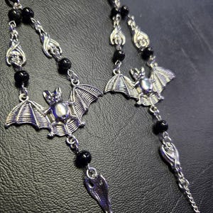 May include: A pair of silver-toned metal chains featuring bat-shaped charms and smooth black spherical beads. The design incorporates detailed bat figures with spread wings, connected by delicate silver links and interspersed with round black beads.