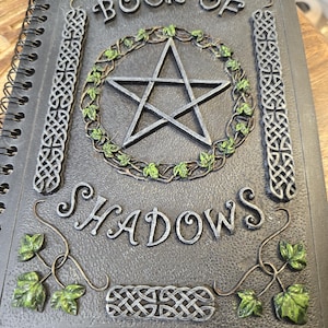 May include: A spiral-bound journal titled "BOOK OF SHADOWS" with a pentagram design. The cover is dark gray with silver accents and green ivy details. The design includes Celtic knot patterns.