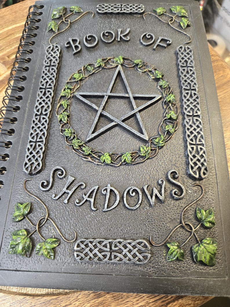 May include: A spiral-bound journal titled "BOOK OF SHADOWS" with a pentagram design. The cover is dark gray with silver accents and green ivy details. The design includes Celtic knot patterns.